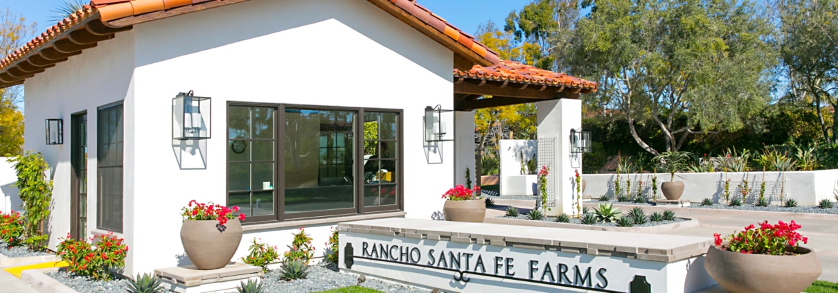 Entrance to Rancho Santa Fe Farms featuring Spanish-style architecture, landscaped gardens, and the community signage set against a clear blue sky.