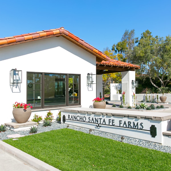 Entrance to Rancho Santa Fe Farms featuring Spanish-style architecture, landscaped gardens, and the community signage set against a clear blue sky.