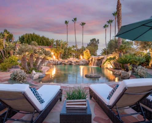 Resort-style backyard with lounge chairs overlooking a lagoon pool with waterfall, fire features, and palm trees at sunset Resort-style backyard with lounge chairs overlooking a lagoon pool with waterfall, fire features, and palm trees at sunset
