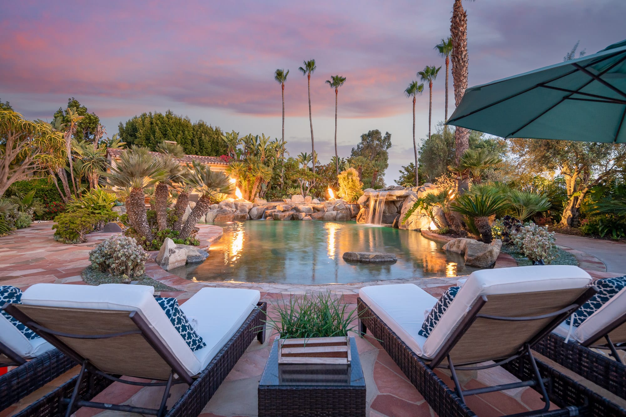 Resort-style backyard with lounge chairs overlooking a lagoon pool with waterfall, fire features, and palm trees at sunset Resort-style backyard with lounge chairs overlooking a lagoon pool with waterfall, fire features, and palm trees at sunset