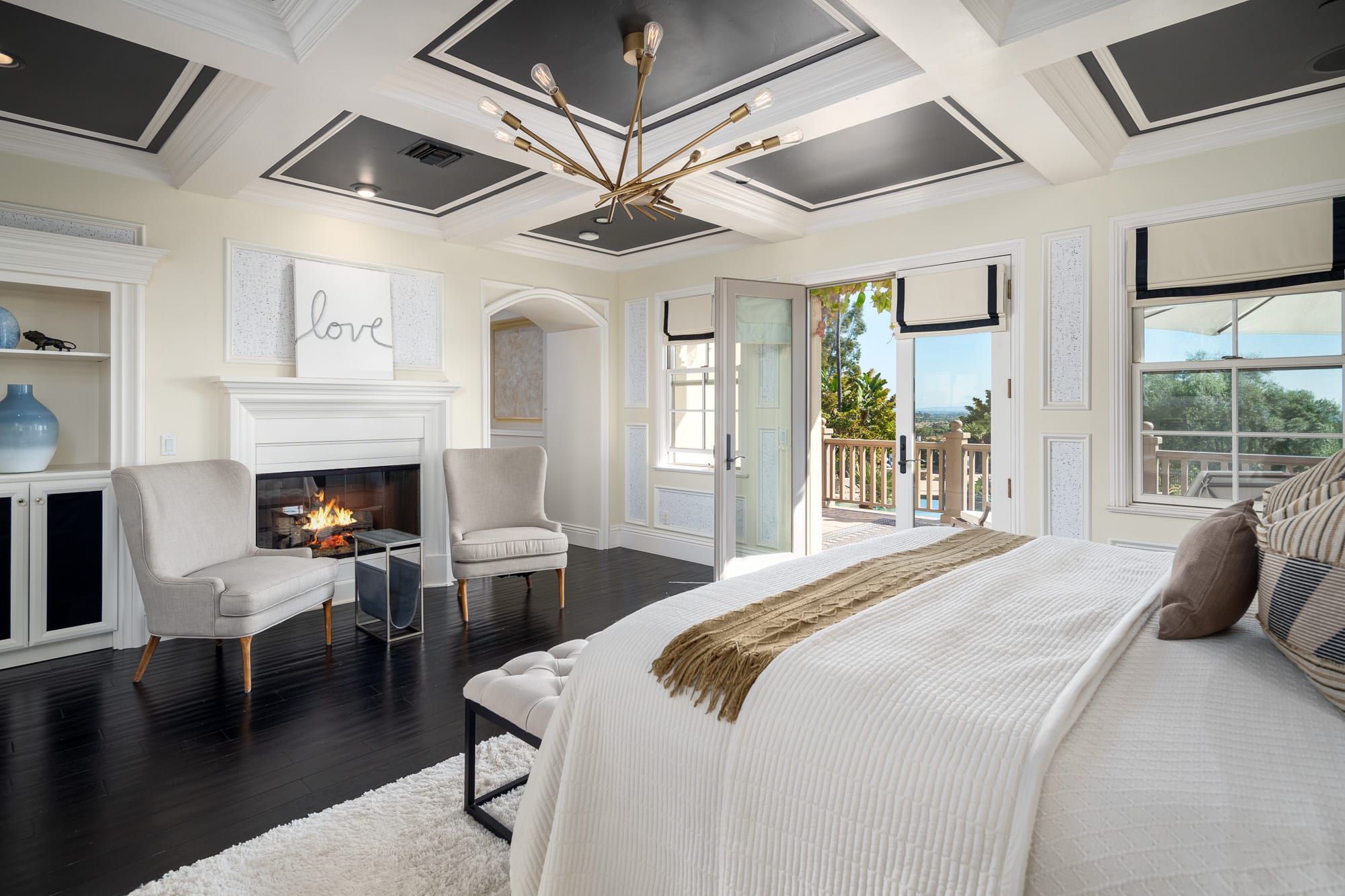 Elegant bedroom with coffered ceiling, statement chandelier, fireplace seating, and French doors opening to a private balcony Elegant bedroom with coffered ceiling, statement chandelier, fireplace seating, and French doors opening to a private balcony