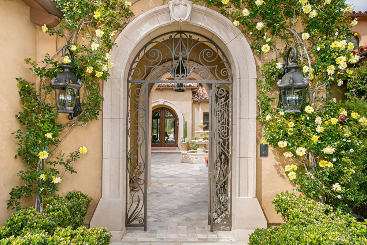 Arched garden entry of a Rancho Santa Fe estate with wrought iron gate, climbing roses, stonework, and landscaped courtyard