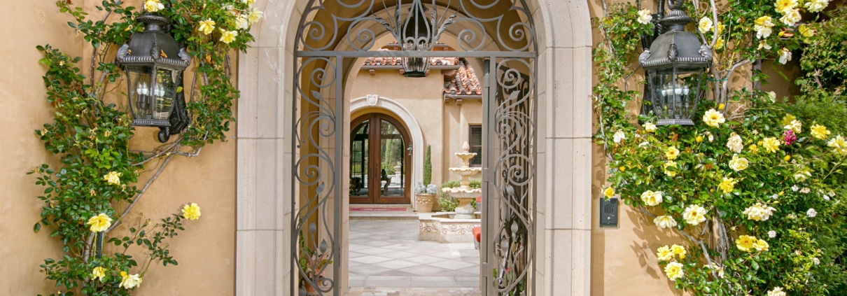 Arched garden entry of a Rancho Santa Fe estate with wrought iron gate, climbing roses, stonework, and landscaped courtyard
