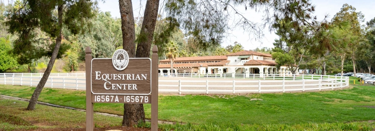 Sign for the Rancho Santa Fe Equestrian Center with paddocks, fencing, and historic buildings in the background