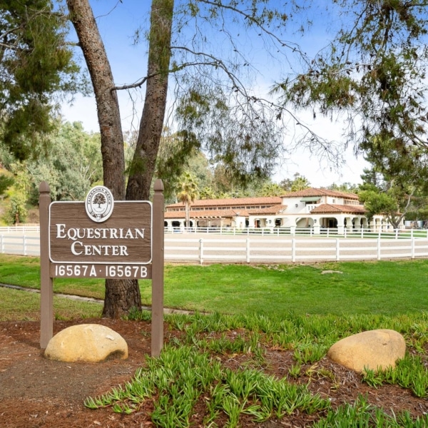 Sign for the Rancho Santa Fe Equestrian Center with paddocks, fencing, and historic buildings in the background