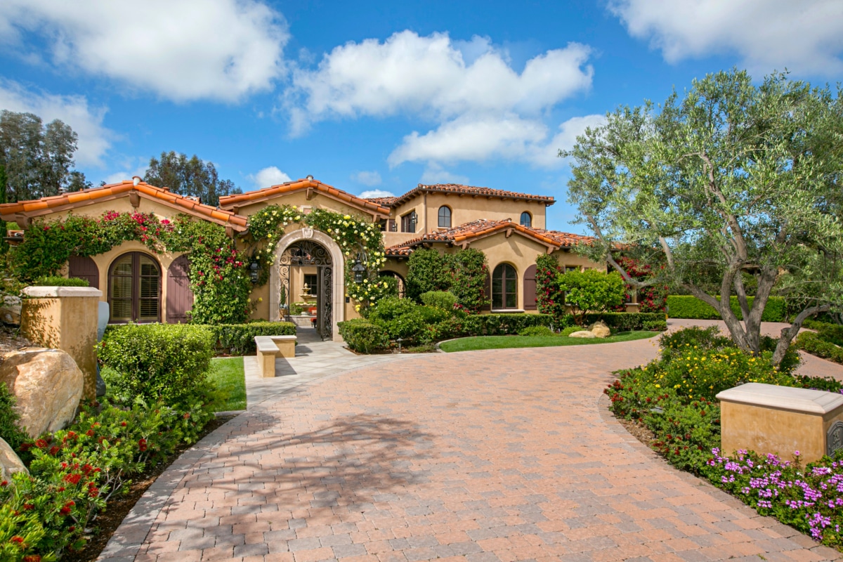 Mediterranean-style estate exterior in Rancho Santa Fe with landscaped gardens, arched entry, and tiled roof under a blue sky