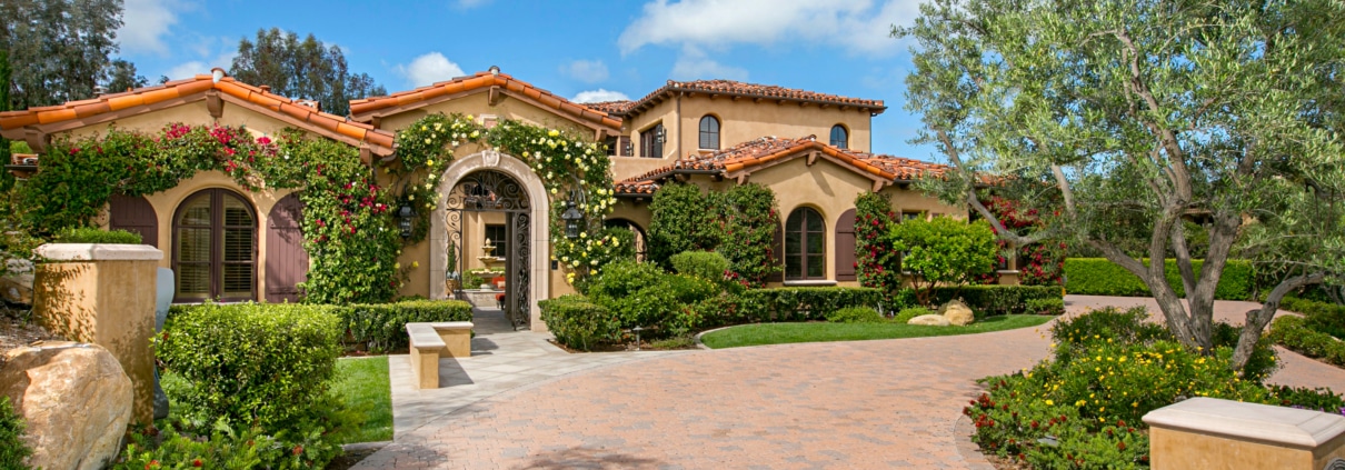 Mediterranean-style estate exterior in Rancho Santa Fe with landscaped gardens, arched entry, and tiled roof under a blue sky