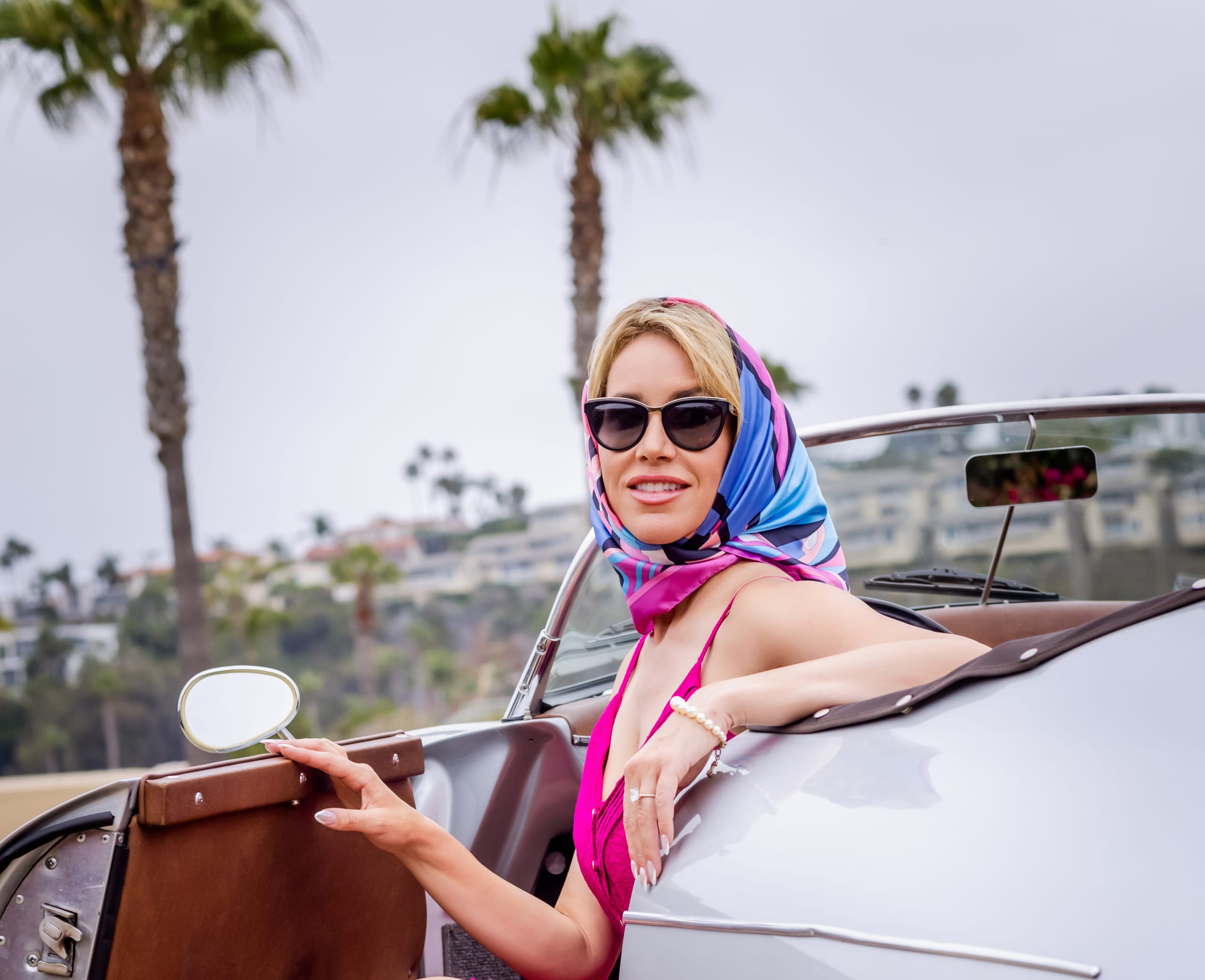 Woman in pink dress and headscarf sitting in a vintage convertible styled for Del Mar Opening Day race day fashion