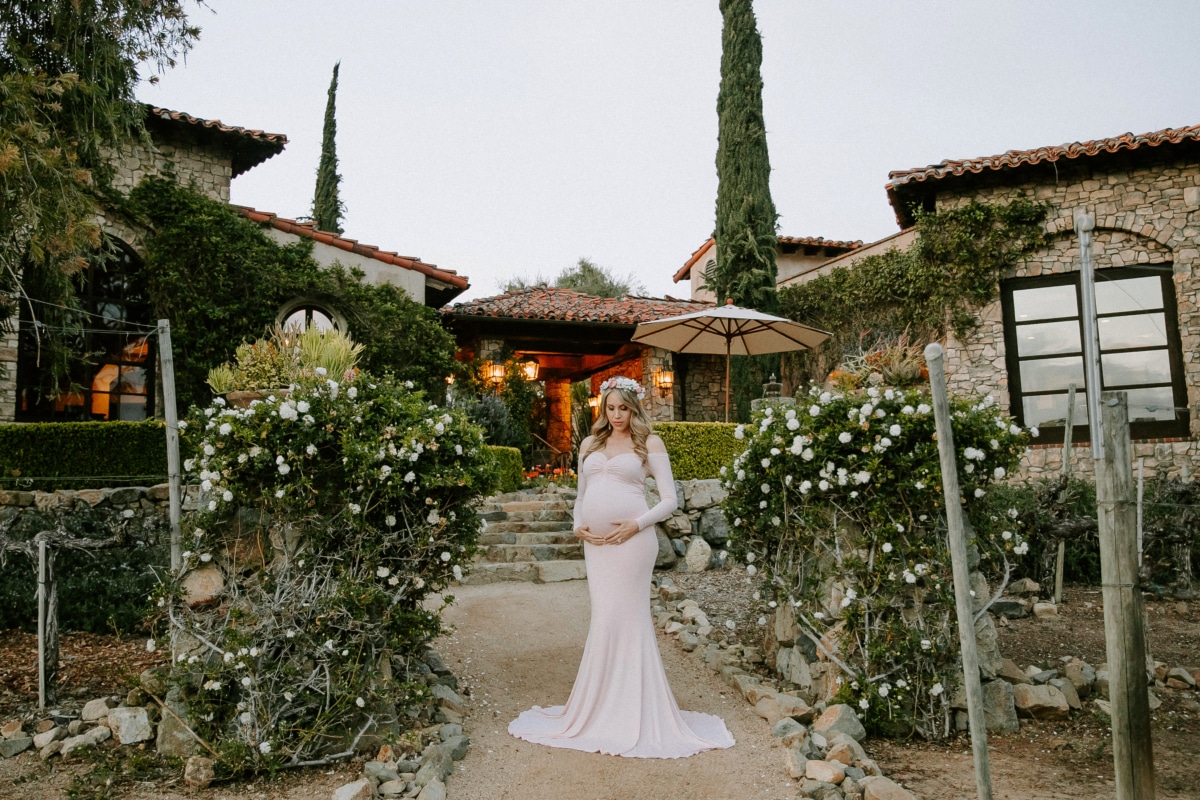 Priscilla at The Bridges, drawn to the light and quiet elegance of the setting Pregnant woman in a soft pink gown standing in front of a luxury estate at The Bridges in Rancho Santa Fe