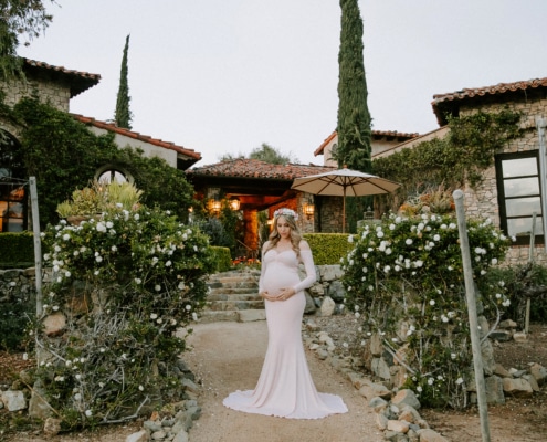 Priscilla at The Bridges, drawn to the light and quiet elegance of the setting Pregnant woman in a soft pink gown standing in front of a luxury estate at The Bridges in Rancho Santa Fe